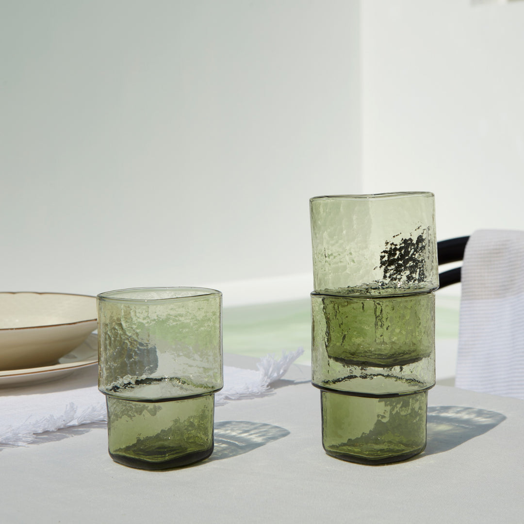 Stackable olive-green handmade drinking glasses with a unique form and hammered texture on an outdoor dining table on a summer day