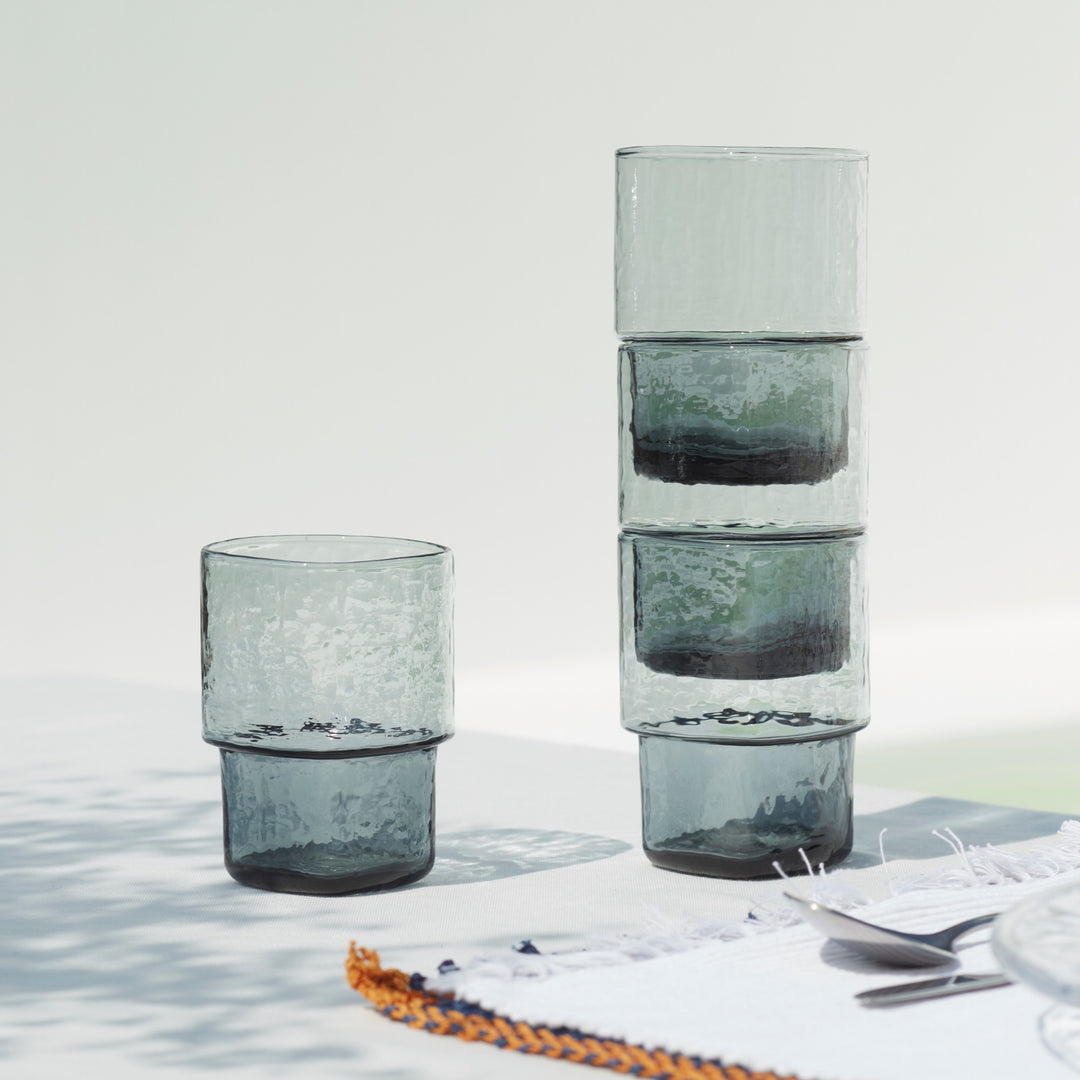 A set of 4 unique, stackable handmade smoke-grey drinking glasses with an unusual shape and hammered texture on a summer table setting 